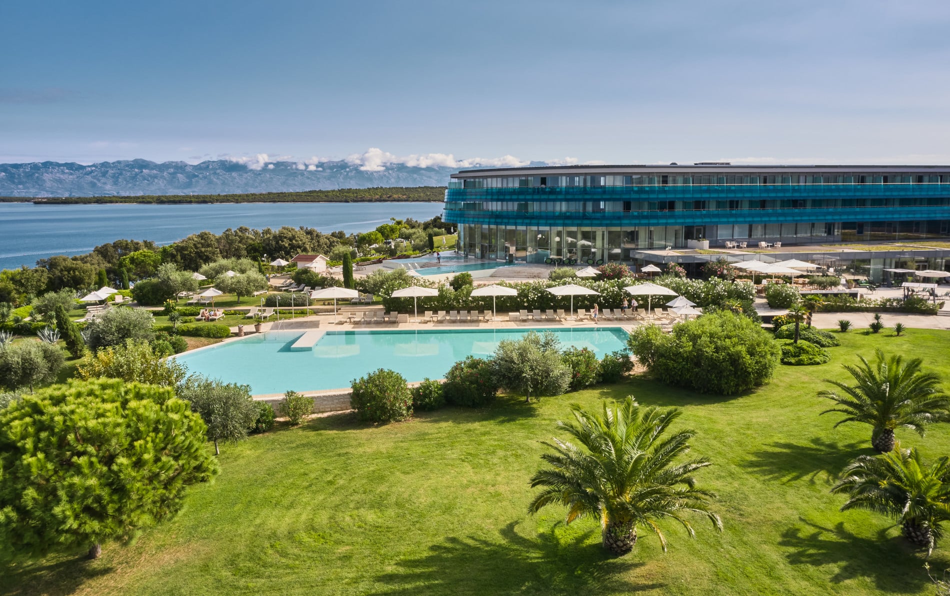Das Falkensteiner Hotel Spa Iadera ist zu sehen, das dirkt am Meer liegt, zudem ist eine Poollandschaft, grüne Wiese und viele Palmen zu sehen.