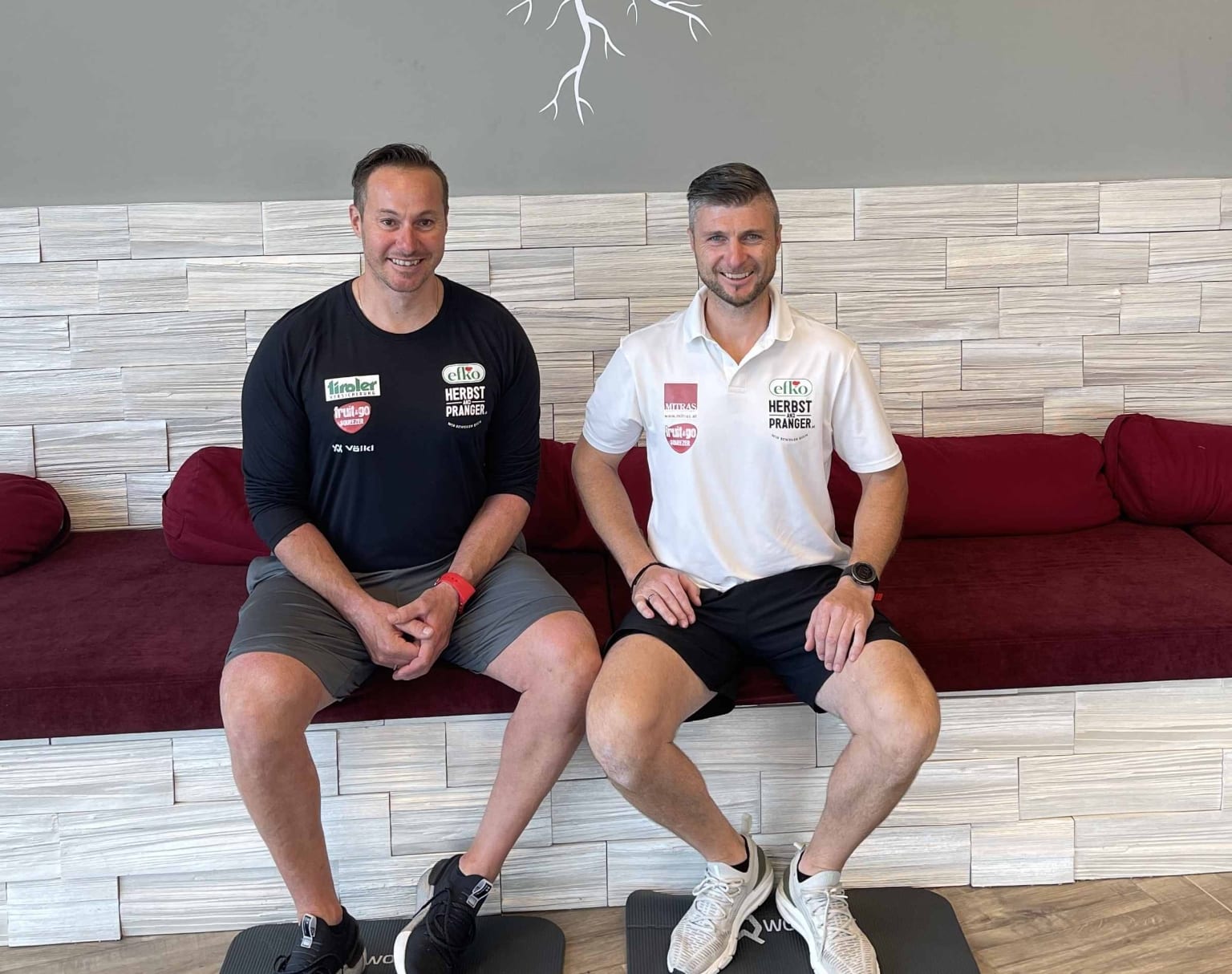 Die beiden Skilegenden Manfred Pranger und Reinfried Herbst sitzen auf einer Bank mit roten Pölstern